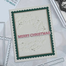 A handmade Christmas greeting card with a scalloped dark green border, an embossed holly pattern, and a red 'MERRY CHRISTMAS' banner, shown on a light gray background with crafting tools.