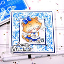 A handmade greeting card featuring a cute orange tabby cat in a blue bow and blue outfit, surrounded by blue floral patterns. The card includes the text 'TRUE beauty is felt in the heart' and is displayed on a white surface with craft tools and rhinestones.