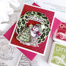 A handmade Christmas card featuring a penguin sitting on a small stool beside a decorated tree, with the text 'Deck the Halls with Lots of Joy!'. The card is layered over a red textured background and pink envelope, with Gina K.