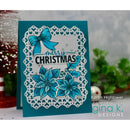 A handmade Christmas greeting card with a teal background, featuring a white scalloped die-cut border, blue poinsettia flowers, a blue bow, and the words 'Merry Christmas' in black script and block lettering. The card is displayed on a tabletop with blurred holiday decorations in the background.
