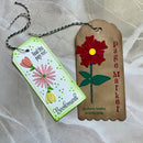 Two handmade paper bookmarks with floral embellishments, displayed on a light beige textured fabric background. One bookmark is white with green dotted borders, featuring pink and red flowers, yellow accents, and the text 'Bookmark' and 'Read this page next.