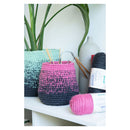 A crocheted basket organizer in pink and dark gray sits on a white shelf next to a mint green and gray crocheted basket, with a pink and white ball of yarn in front. A white ceramic vase and a potted plant are in the background.