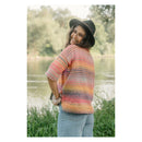 A woman wearing a colorful striped knit sweater, blue jeans, and a black hat stands outdoors by a river with trees in the background.