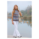 A woman stands by a calm lake wearing a sleeveless, high-neck knit top with a blue, orange, and brown striped pattern, white pants, and carrying a beige shoulder bag.