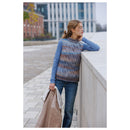 A woman stands outdoors leaning on a light stone wall, wearing a sleeveless knit sweater vest with a blue, brown, and beige striped pattern over a light blue long-sleeve shirt and dark blue jeans. She is holding a tan jacket.