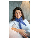 A smiling woman with curly brown hair wearing a light blue shirt and a textured blue knit scarf, seated indoors against a gray wall with a warm lamp in the background.