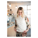 A woman smiling in a cozy indoor shop setting, wearing a cream cable-knit sweater vest over a long-sleeve paisley blouse.