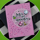 A square pink card with a circular clear sticker reading 'Let's get spookty' and small spooky illustrations, placed on a black-and-white checkered plate over a bright green surface.