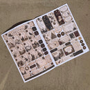 An open booklet or album page filled with vintage Christmas-themed labels, stickers, and decorative ephemera, photographed from above on a textured tan fabric background.