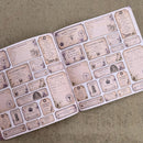 An open planner or notebook spread showing multiple vintage-style label sheets with gothic and occult-themed text, skulls, botanical illustrations, and ciphers, laid on a textured beige fabric background.