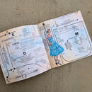 An open vintage sewing pattern book laid flat on a textured beige fabric surface. The pages are aged cream with blue watercolor washes, black printed text and measurements, and a central illustration of a woman in a blue dress.