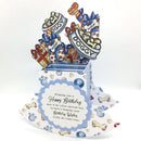 A 3D pop-up dog birthday card with a blue and white striped base decorated with bone and paw print motifs. The card features a central blue scalloped label with a message for a happy birthday, and rising from the base are cartoon gifts wrapped in red, gold, and blue ribbon, topped with treats and banners reading 'DOGGY' and 'WOOF'.