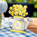 A decorative watering can-shaped card or gift holder filled with yellow daffodils, placed on a blue-and-white checkered tablecloth beside a glass vase of blue flowers. The front has a round label reading “Love & Birthday Wishes.