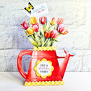 A red watering can shaped vase filled with colorful tulips, featuring a yellow scalloped trim, a flower pot emblem with 'Love & Birthday Wishes', and a butterfly tag that reads 'Especially for you!'.