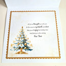 An open greeting card with a festive New Year design. The card features a watercolor-style decorated Christmas tree on the left and elegant black script text on the right that reads, 'All of our thoughts are with you.