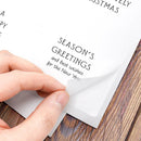 A close-up of a hand peeling back a transparent protective film from a white paper holiday greeting card placed on a wooden surface. The card shows printed text including 'SEASON'S GREETINGS' and partial 'MERRY CHRISTMAS'.