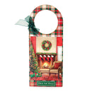 A Christmas-themed wooden hanging sign with a hanger loop at the top, featuring a red and green plaid border, a green ribbon bow, and a central printed holiday scene of a decorated fireplace, wreath, tree, stockings, and a chair. The sign reads 'Deck the Halls' at the bottom.