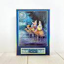 A vertical art print or card featuring the Three Wise Men under a full moon, with a holographic iridescent blue border and a small matching strip at the bottom, displayed on a light wood surface against a white wall.