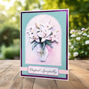 A handmade sympathy greeting card featuring white lilies in a glass vase, set against a teal and purple layered background with the text 'Deepest Sympathy' on a wooden table with a blurred garden background.