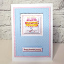 A handmade birthday card featuring a layered blue and pink paper design with a central illustration of a yellow cake and the text 'Happy Birthday To You' at the bottom, displayed against a white brick wall background.