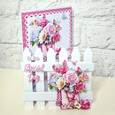 A handmade greeting card displayed on a white picket fence stand against a white brick wall. The card features a pink and white patterned border, a bouquet of pink and blue flowers, butterflies, and the text 'On Your Special Day'.
