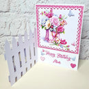 A handmade 3D pop-up birthday card for 'Mum' featuring a pink and white floral bouquet in a checkered vase, a white picket fence on the left, and 'Happy Birthday Mum' written in pink script on a white background.