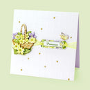 A handmade greeting card with a white embossed grid background, decorated with a 3D floral basket of yellow and purple flowers, a green and yellow 'Primrose' label, a butterfly embellishment, and small gold rhinestones, shown on a pale green background.
