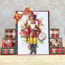 A festive handmade Christmas card featuring a detailed nutcracker illustration with red and gold attire, white beard, and holly accents, displayed on a wooden surface with matching plaid gift boxes and small round cards.