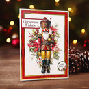A handmade Christmas greeting card featuring a detailed nutcracker illustration with red and gold armor, surrounded by holly and red berries. The card has a red patterned border and the text 'Christmas Wishes' on the top left, standing on a wooden surface with pinecones and blurred holiday lights in the background.