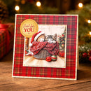 A square Christmas greeting card standing on a wooden surface, featuring a red plaid border and a central illustration of two kittens wrapped in a red plaid blanket inside a basket, with a gold circular badge that reads 'Just for YOU'. The background shows a softly blurred festive holiday scene with lights and greenery.