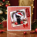 A square Christmas greeting card with red and white vertical stripes, featuring a central photo of a black cat wearing a Santa hat, surrounded by holiday garlands and ornaments. The card is displayed on a wooden surface with festive decorations and blurred Christmas lights in the background.
