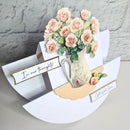 A 3D pop-up greeting card featuring a bouquet of pale pink roses in a vintage floral pitcher, set against white curved paper wings with printed phrases: 'In our thoughts' and 'GOT ALL YOURS'.