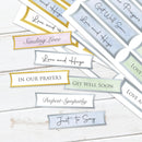 A set of printed sympathy and support message labels or stickers arranged on a white wood surface. The labels have soft pastel colors including pink, gold, blue, green, and gray, with handwritten-style text such as 'Love and Hugs,' 'Sending Love,' 'In Our Prayers,' 'Get Well Soon,' 'Deepest Sympathy,' and 'Just to Say.
