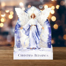 A decorative Christmas card or display stand featuring an angel figure with white wings and a blue robe, standing on a base printed with the words 'CHRISTMAS BLESSINGS.' The background shows warm blurred holiday lights and a wooden surface.