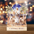 A 3D papercraft Christmas nativity scene display featuring a wise man in white robes standing with three sheep, flanked by palm trees under a starry night sky, set on a wooden base printed with 'CHRISTMAS WISHES'.