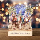 Pop-up Christmas card with three wise men and camels on a wooden surface with blurred lights in the background.