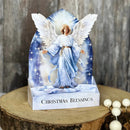 A decorative Christmas angel figurine displayed on a printed arched stand with the words 'CHRISTMAS BLESSINGS' on the base, set on a rustic wood slice with white beads in the foreground.