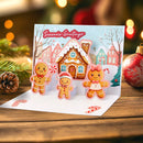 A festive 3D pop-up Christmas greeting card displayed on a wooden surface. The card features three gingerbread men and a gingerbread house with the text 'Season's Greetings' on a snowy hillside, surrounded by holiday decorations like pine branches, a pinecone, and a red ornament in a warm, blurred background.