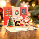 A 3D pop-up Christmas greeting card displayed on a wooden table. The card features a festive living room scene with a decorated Christmas tree, a fireplace with stockings and a burning fire, a framed picture, and a cutout figure of Santa Claus holding a gift bag.