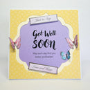 A square greeting card with a yellow patterned border and a central lavender scalloped panel that reads 'Get Well Soon' with smaller text below. Decorative butterfly illustrations appear on the card, along with small ribbon-style labels reading 'Just to Say' and 'Love and Kugs.