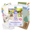 A craft kit package labeled 'Handy Hobby Aprons Card Making Kit' surrounded by included craft supplies, including patterned cardstock, envelopes, scissors, and small decorative components, arranged on a white background.