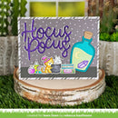 A handmade card or gift tag featuring a purple glittery 'Hocus Pooh' script, a teal potion bottle, a small yellow cat in a witch hat, and other occult-themed illustrations, displayed on a rustic birch log slice with grass and garden decor in the background.