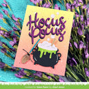 A Halloween-themed handmade card featuring the words 'Hocus Pocus' in glittery purple script above a black cauldron with green slime, white foam, and a broom. The card is styled on a turquoise fabric surrounded by purple flowers.