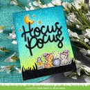 A handmade square card featuring the words 'Hocus Pocus' in black glittery script over a turquoise-to-yellow gradient background, with a crescent moon, stars, a cauldron, and two cartoon cats wearing purple hats. The card is styled on a textured teal surface with white flowers around it.