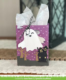 A Halloween-themed paper gift bag with a purple polka-dot background, a white ghost cutout, a black haunted house, and twine bow, standing on a textured surface.