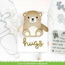 A handmade greeting card featuring a cute layered teddy bear with a gold foil 'hugs' script on a white background, surrounded by crafting tools and a clear stamp sheet.