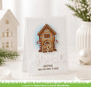 A handmade Christmas greeting card with a scalloped white border, featuring a gingerbread house design with two dogs and the text 'Christmas from our family to yours' and 'Sweet'. The card is displayed on a tabletop with holiday props including a small white house, greenery, and a white vase.