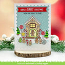 A handmade Christmas greeting card featuring a gingerbread house illustration with the text 'HAVE A SWEET CHRISTMAS FROM OUR FAMILY TO YOURS' on a scalloped white border. The card is displayed on a wood slice stand with blurred holiday greenery and red background bokeh.