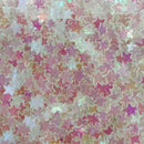 A close-up of many small star-shaped embellishments or confetti in pastel pink, lavender, peach, and iridescent white tones, filling the frame.