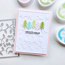 A handmade greeting card with a white embossed background, a pink border, and four colorful pine trees above the words 'DECK THE HALLS'. The card is styled on a white surface with crafting tools, including a metal die and small ink pads.